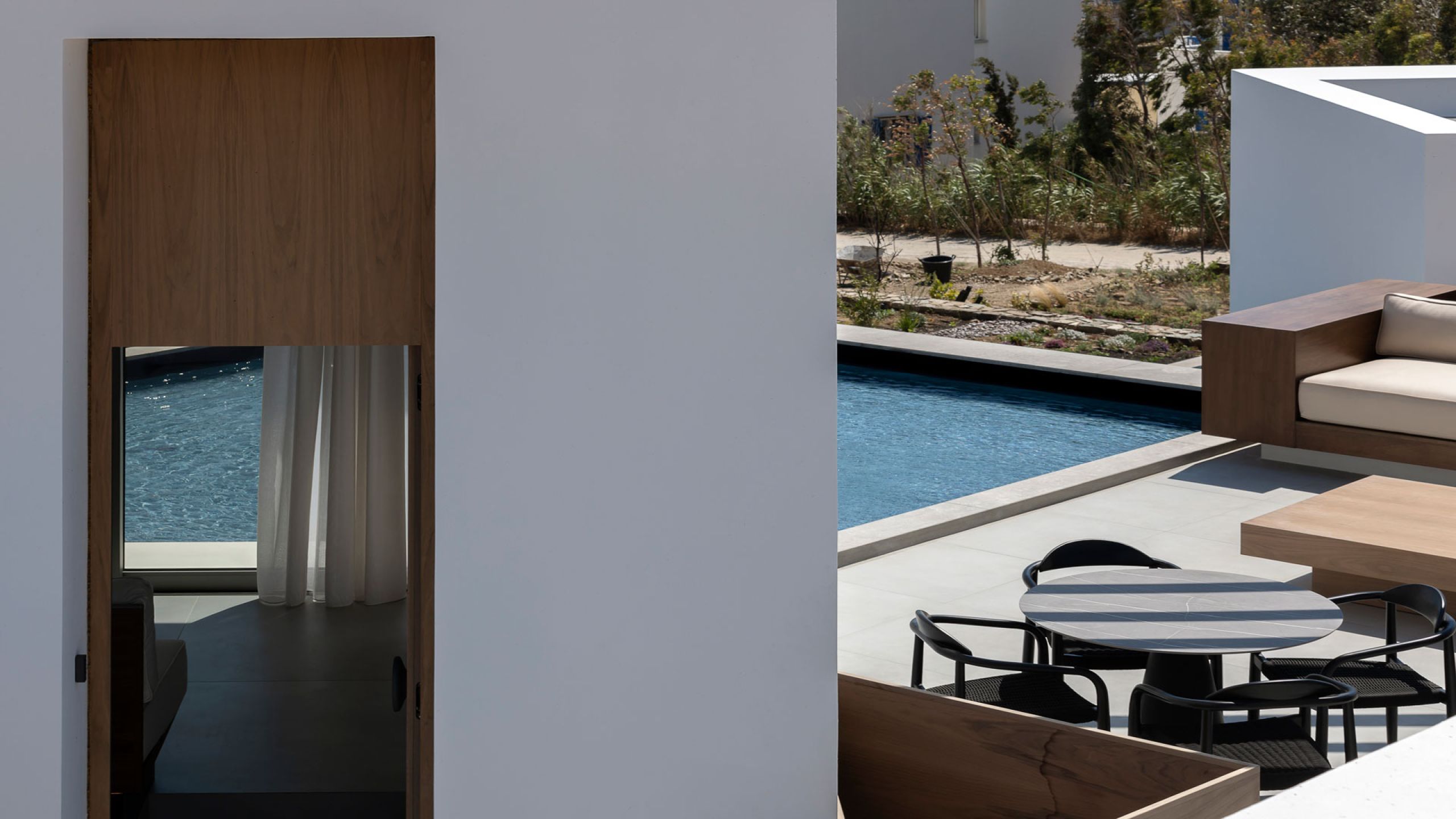 Pnoes Tinos terrace: round dining table with black chairs and a timber lounge beside a pool, with a doorway glimpsing a curtained room