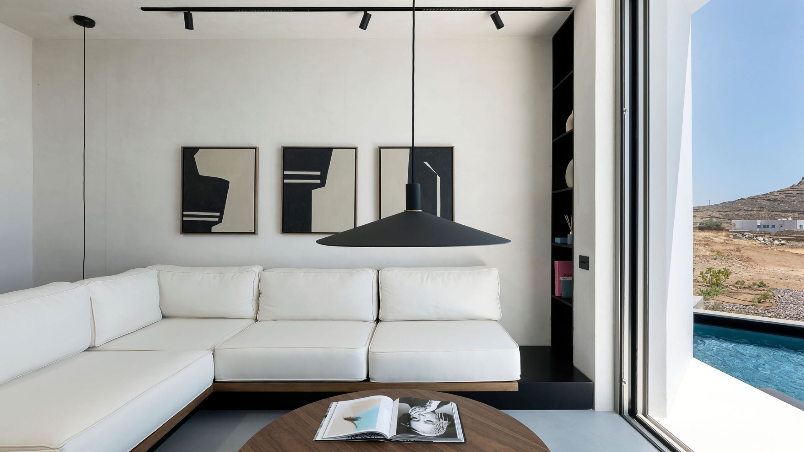 Pnoes Tinos living room: white corner sofa, walnut coffee table, three framed abstract prints and a window onto the pool and hills