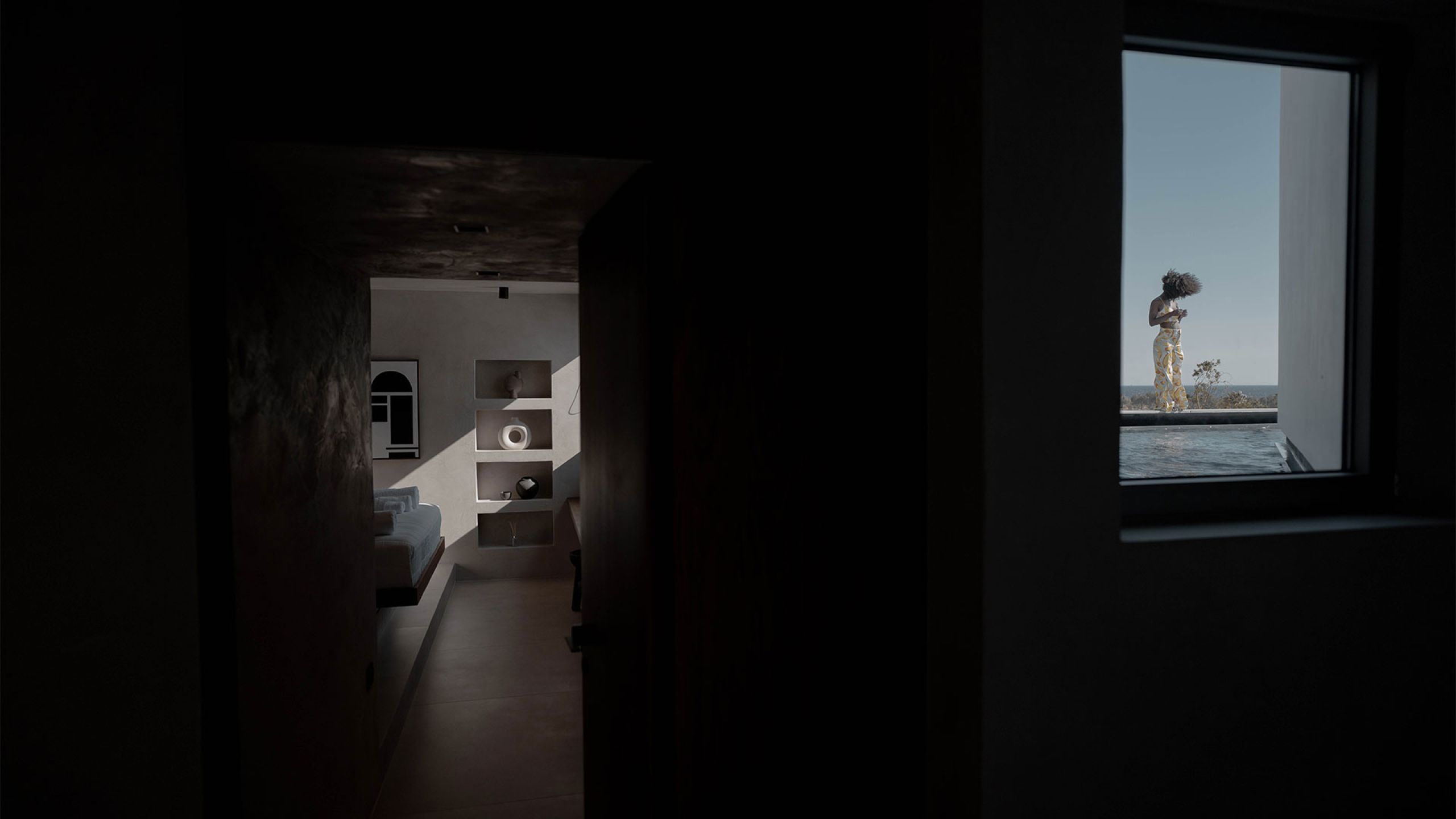 Pnoes Tinos seen from a dark interior: a hallway to a bright bedroom, and a window framing a person beside the pool and sea