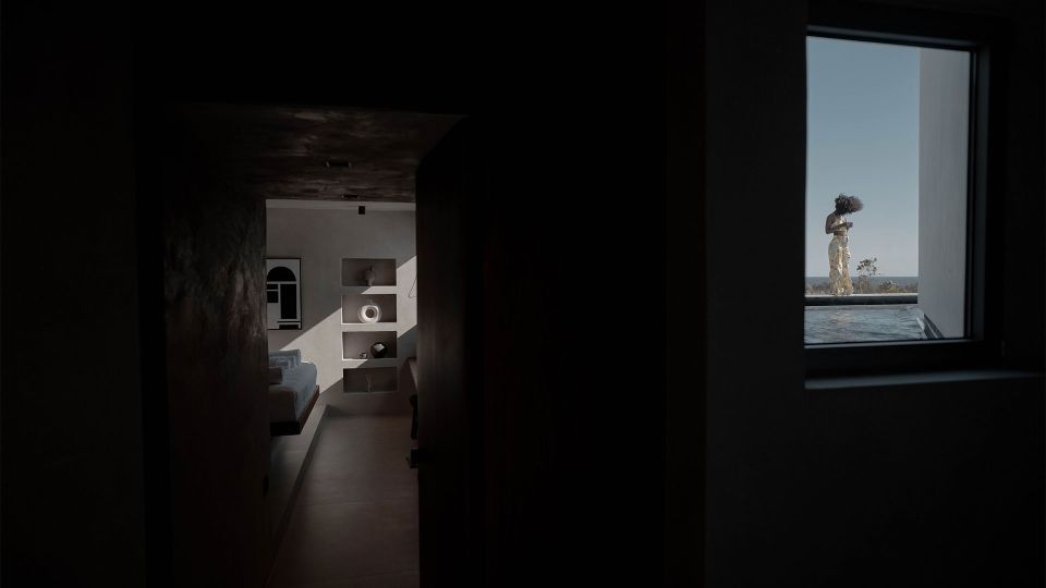 Pnoes Tinos seen from a dark interior: a hallway to a bright bedroom, and a window framing a person beside the pool and sea