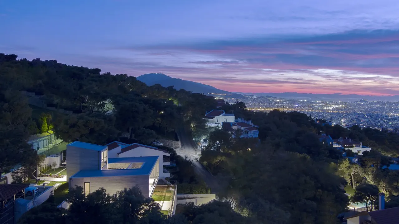Residence in Penteli: the house lit at dusk on a wooded hillside above the lights of Athens